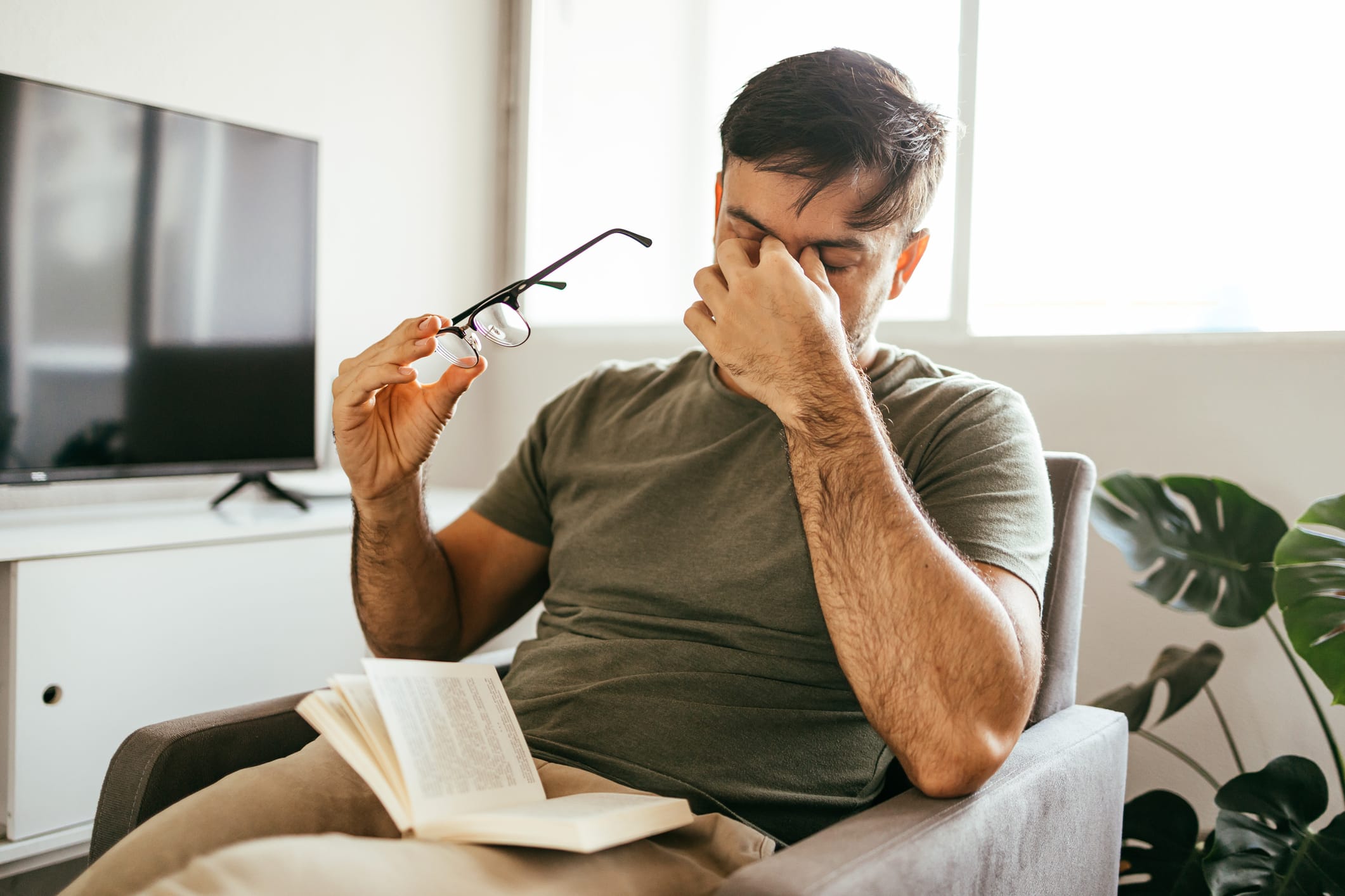 Tired man takes off his glasses and rubs his eyes while reading a book at home.