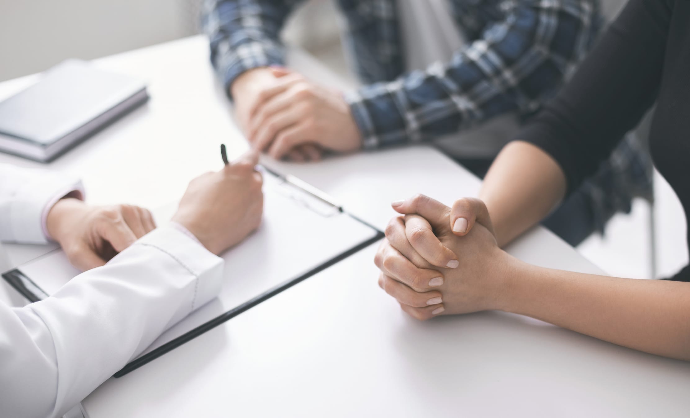 Hands of man and woman resting on a table while doctor writes on a clipboard