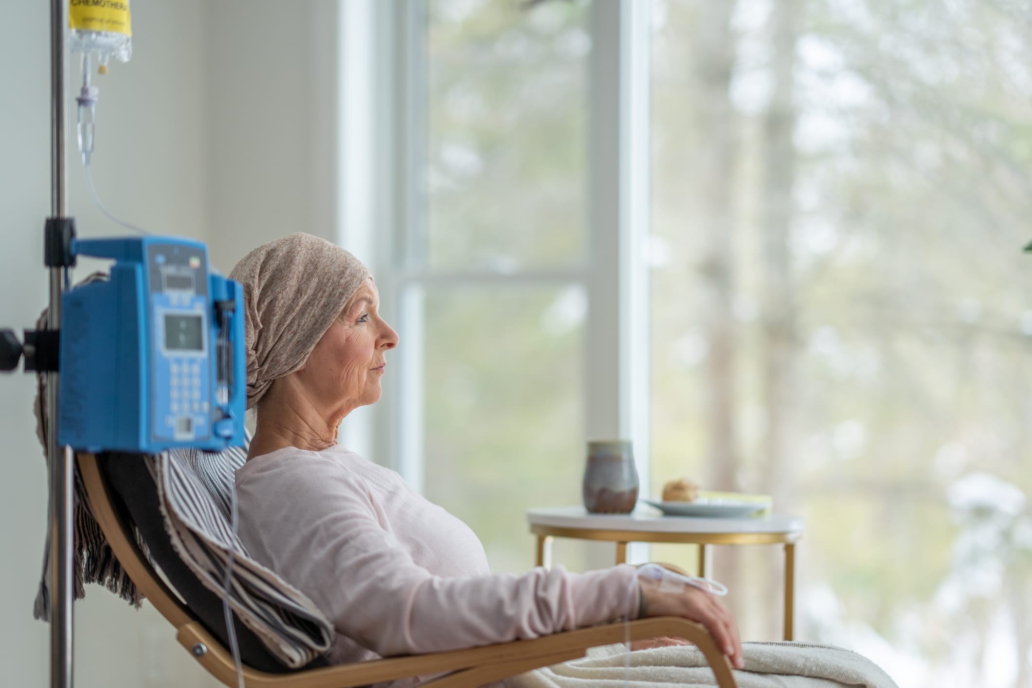 A senior woman sits in the comfort of her own home as she receives her chemotherapy treatment. She is dressed comfortably and wearing a headscarf to keep her warm.