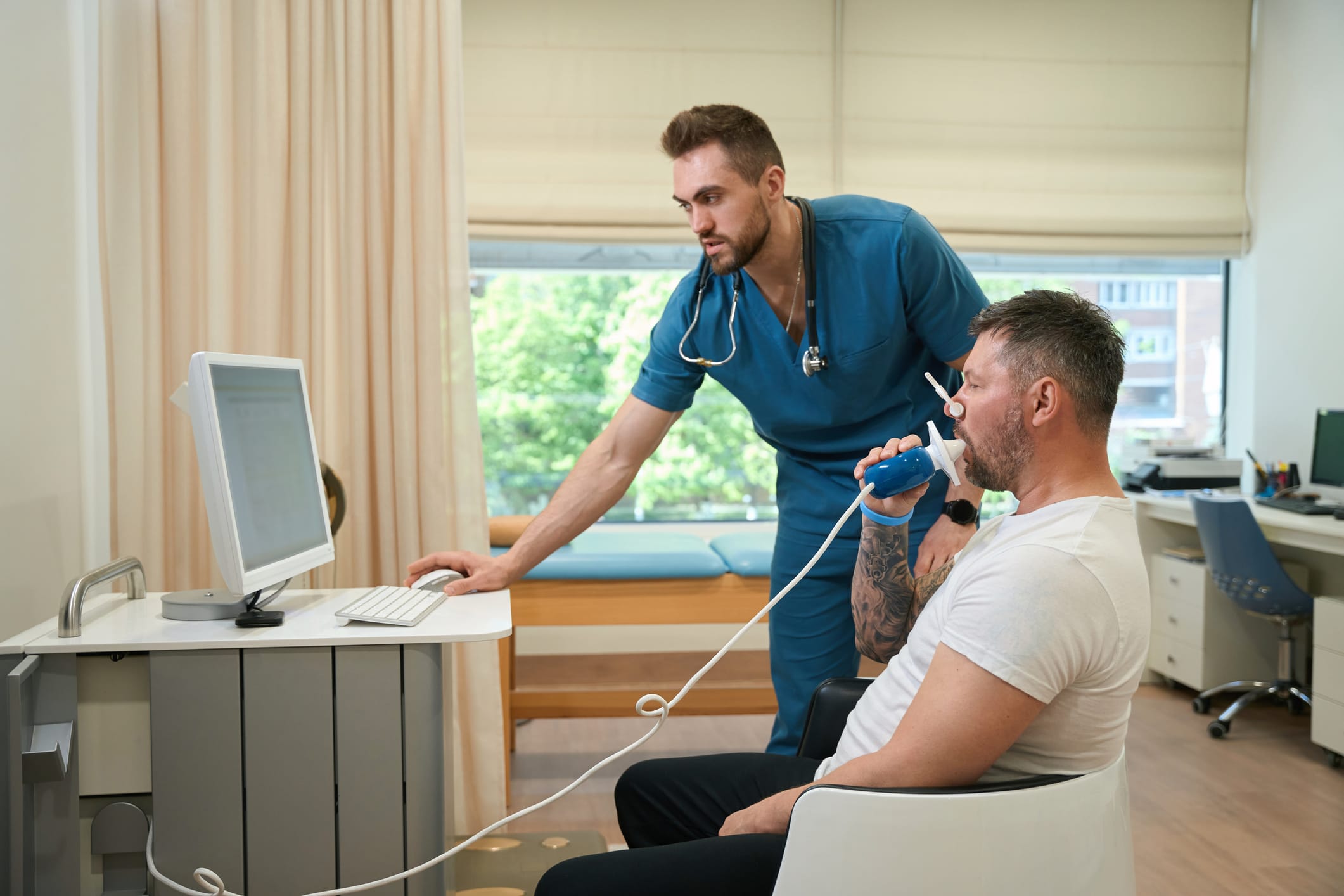 Patient with nose clip breathing into spirometer mouthpiece while pulmonologist monitoring his lung function on computer screen